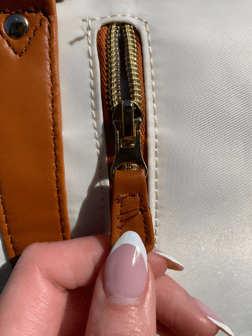 Close-up of a zipper on a beige bag with a brown pull tab and stylish nails holding it.